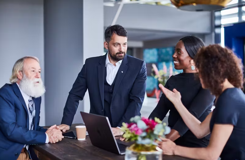 Four diverse workers gathered around a table and laptop to illustrate Microsoft Places + ISS Workplace Experience App