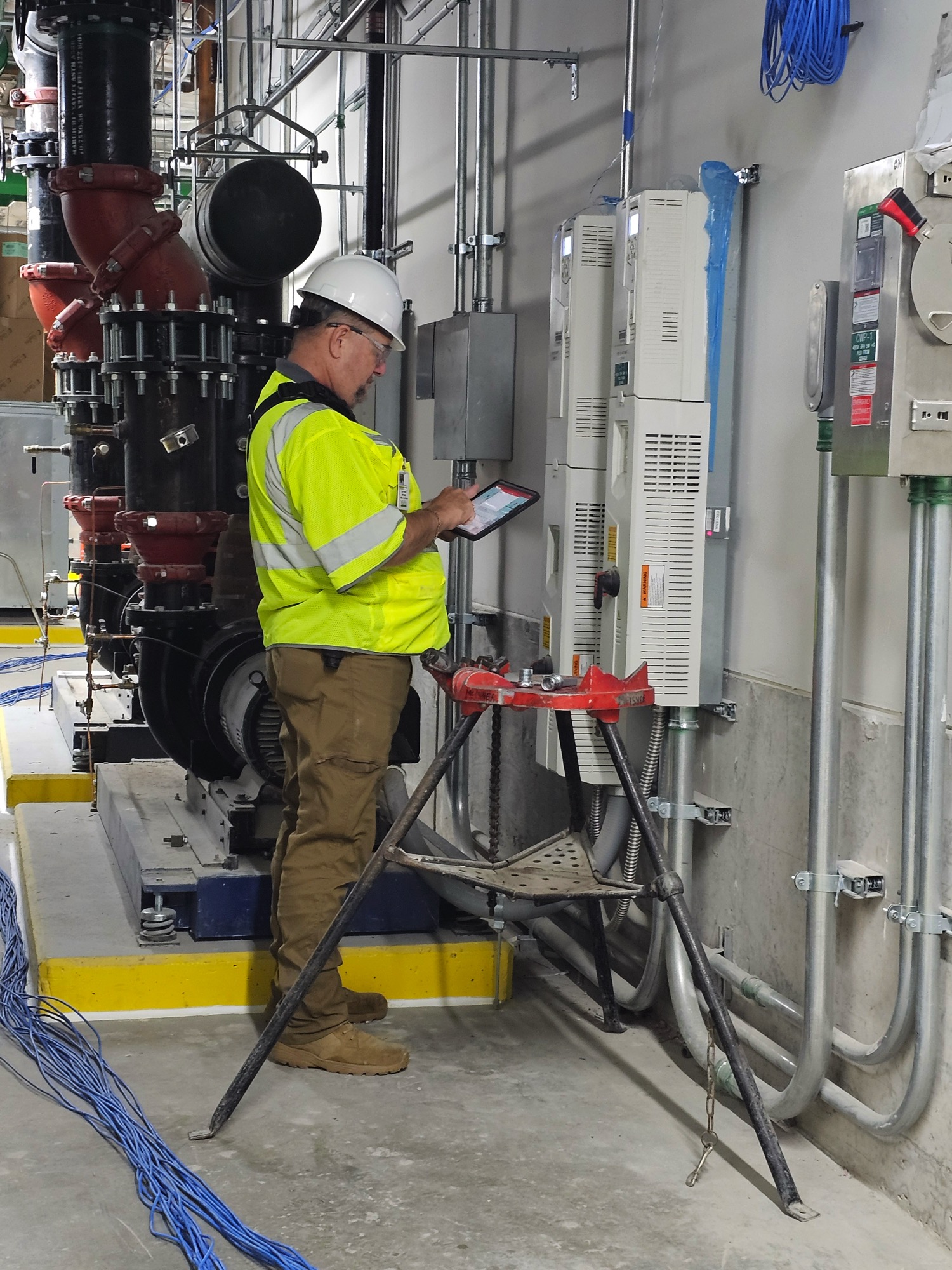 A technician in a yellow safety vest and hard hat using his tablet and checking equipment on a wall