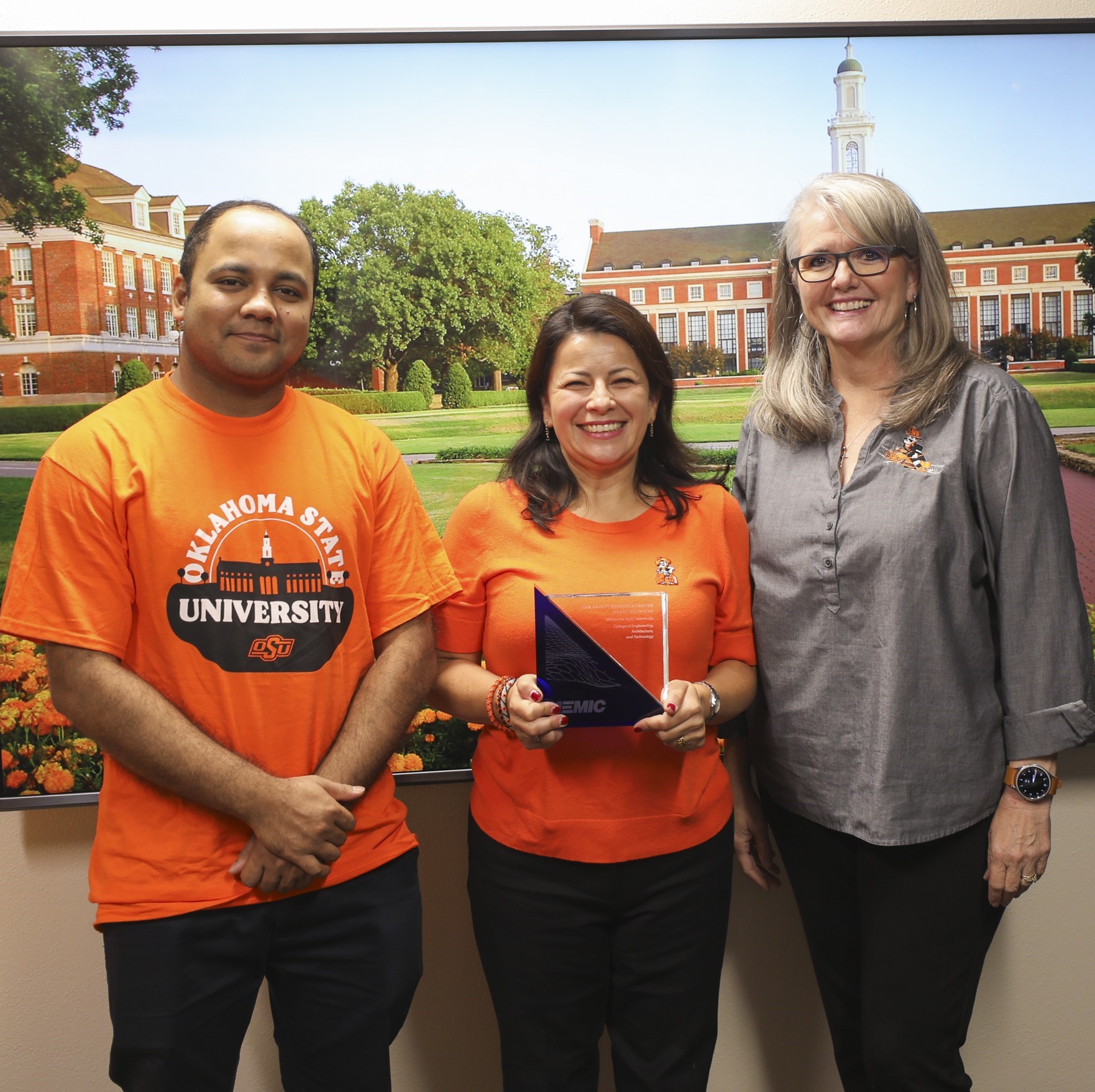 MEMIC image of 3 OSU spiritwear-clad grant recipients holding an award, in front of a screen of OSU campus